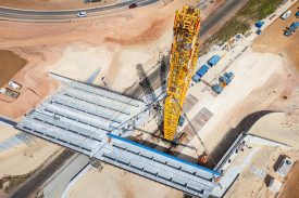 A high-angle view of an industrial bridge-building project featuring a massive yellow crane. The construction site includes various materials and equipment systematically arranged, emphasizing urban infrastructure progress. The scene portrays engineering precision, construction development, and industrial work in progress.